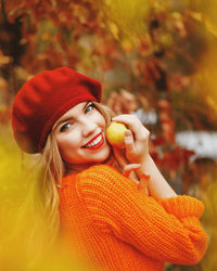 Woman in orange sweater and red beanie holding an apple amidst autumn foliage