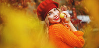 Woman in orange sweater and red beret holding an apple amidst autumn foliage